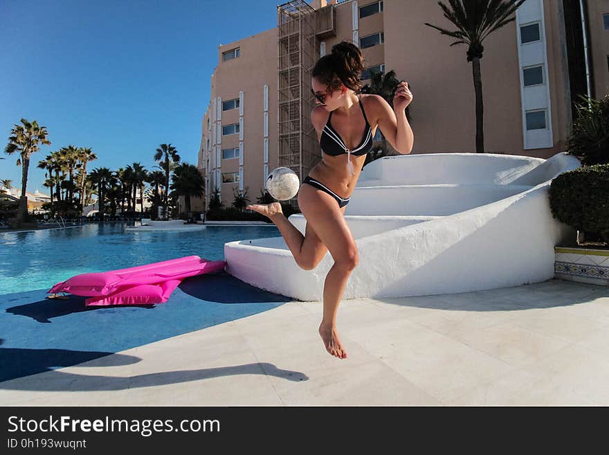 Young attractive woman in a bikini beside a clear blue swimming pool kicking a white football, hotel and trees in the background. Young attractive woman in a bikini beside a clear blue swimming pool kicking a white football, hotel and trees in the background.