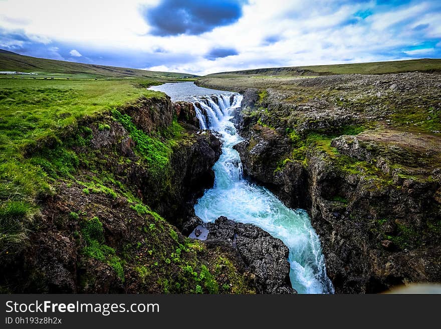 Waterfall over rocky cliffs through green fields on sunny day.