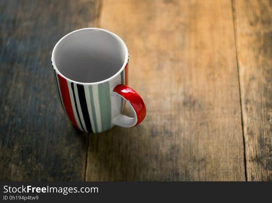 Empty mug with green, black, white and red vertical stripes, on a table made of wooden planking partly (sanded) bleached. Empty mug with green, black, white and red vertical stripes, on a table made of wooden planking partly (sanded) bleached.