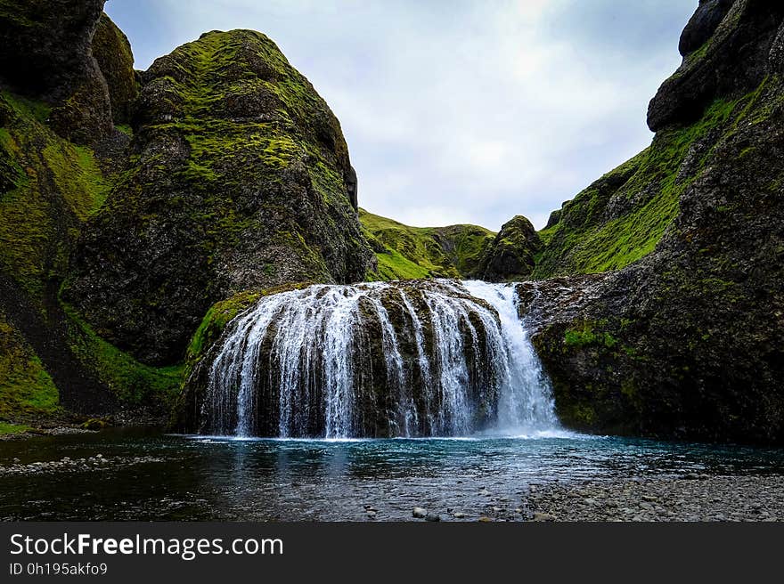 Waterfalls over green cliffs into lake or stream with cloudy skies on sunny day.