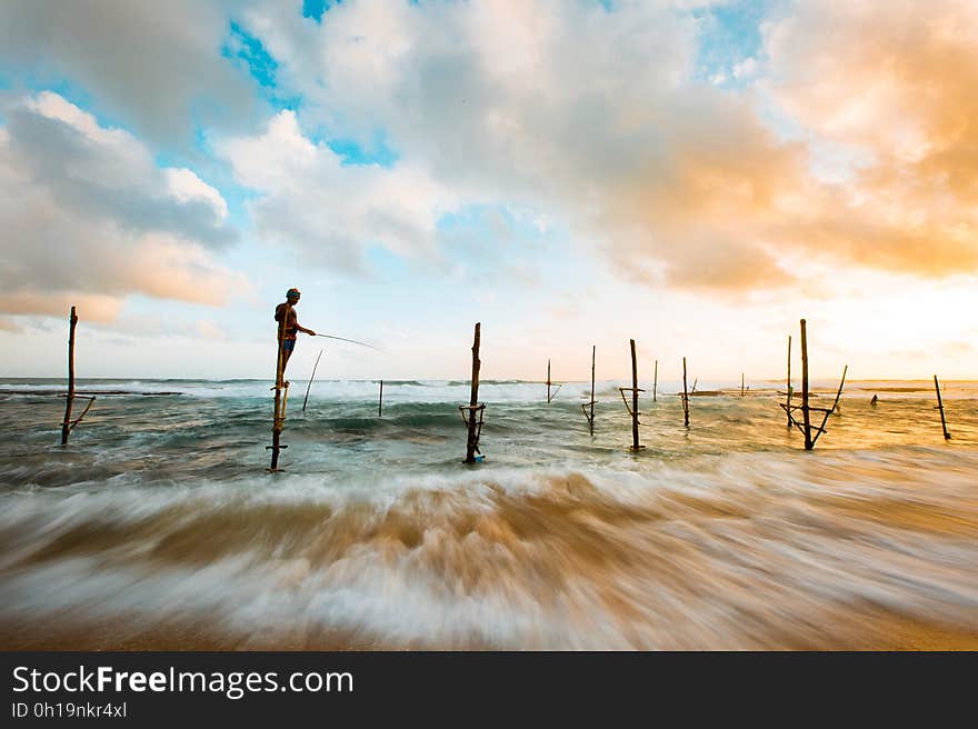 Man Standing in Wood in the Middle of the Ocean While Fishing Under Blue and White Sky during Day Time