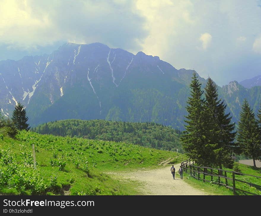 2 Person Walking Beside Grass Field Near Mountain