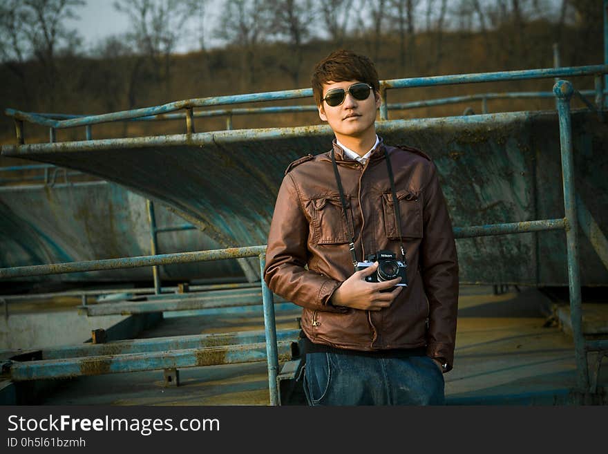 Man in Brown Leather Jacket Wearing Black Framed Sunglasses Holding Black Dslr Camera Standing in Front of Black Steel Bar during Daytime