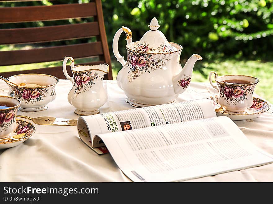 White and Pink Floral Ceramic Tea Set on White Textile Covered Table Beside White and Black Printed Book