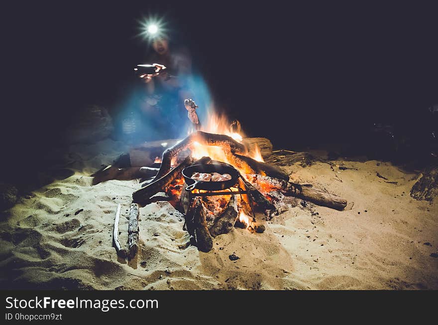 Person wearing headlamp sitting next to campfire on beach with skillet of cooking food. Person wearing headlamp sitting next to campfire on beach with skillet of cooking food.