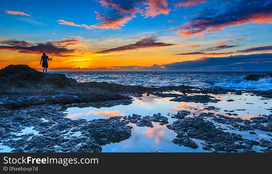 Dramatic orange and pink sunset with lone fisherman seen in silhouette using rod and line fishing from rocks on the seashore of Taiwan. Dramatic orange and pink sunset with lone fisherman seen in silhouette using rod and line fishing from rocks on the seashore of Taiwan.