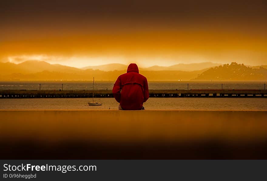 Person Wearing Red Hoodie Sitting In Front of Body of Water