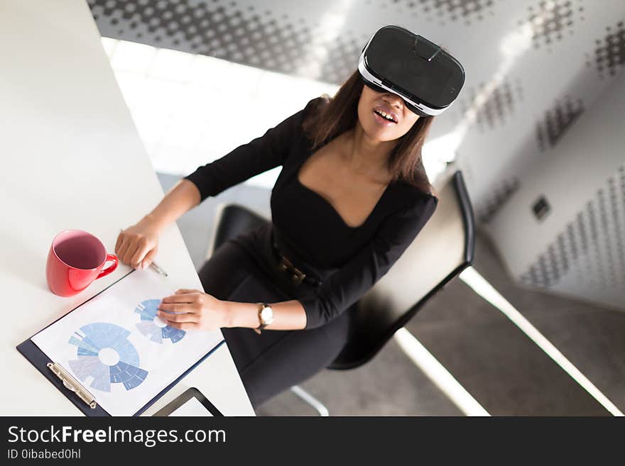 Beauty young brunette afroamerican business lady in black business suit, glasses and costly watch sit at the table in the office, red cup on the table, sit in virtual reality glasses, turned picture. Beauty young brunette afroamerican business lady in black business suit, glasses and costly watch sit at the table in the office, red cup on the table, sit in virtual reality glasses, turned picture