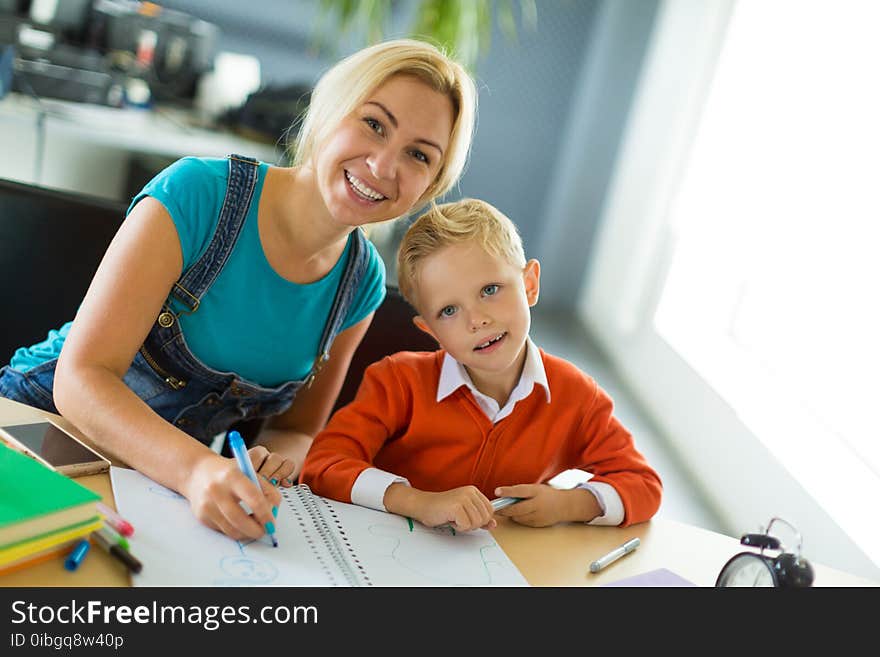 Cute Boy And His Mom Sit At The Desk In The Office And Draw