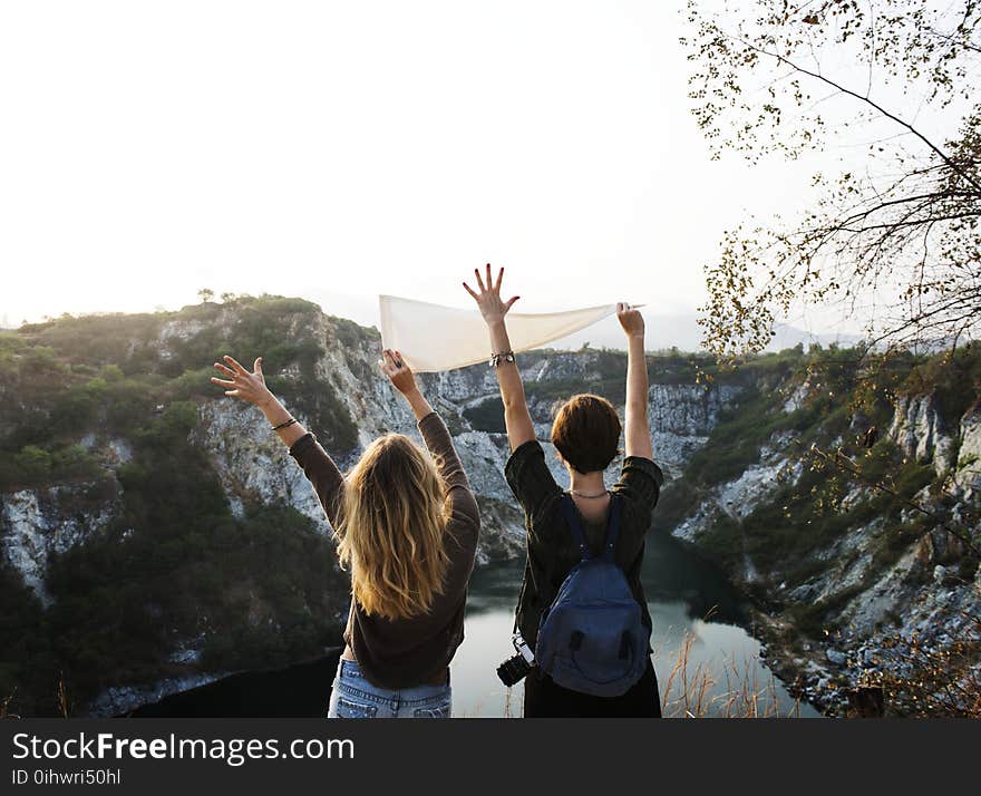 Man Holding White Pennant Beside Girl Raising Two Hands in Front of Lake Surrounded by Mountains