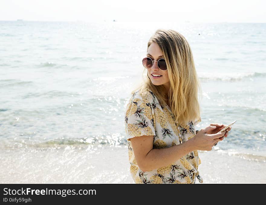 Woman in Black Yellow and White Floral Button-up Shirt Holding Smartphone Wearing Aviator Sunglasses Near Body of Water