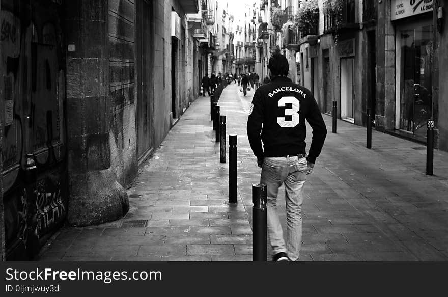 Grayscale Photo of Man Wearing Long-sleeved Shirt and Pants Walking on Concrete Pathway Near Building