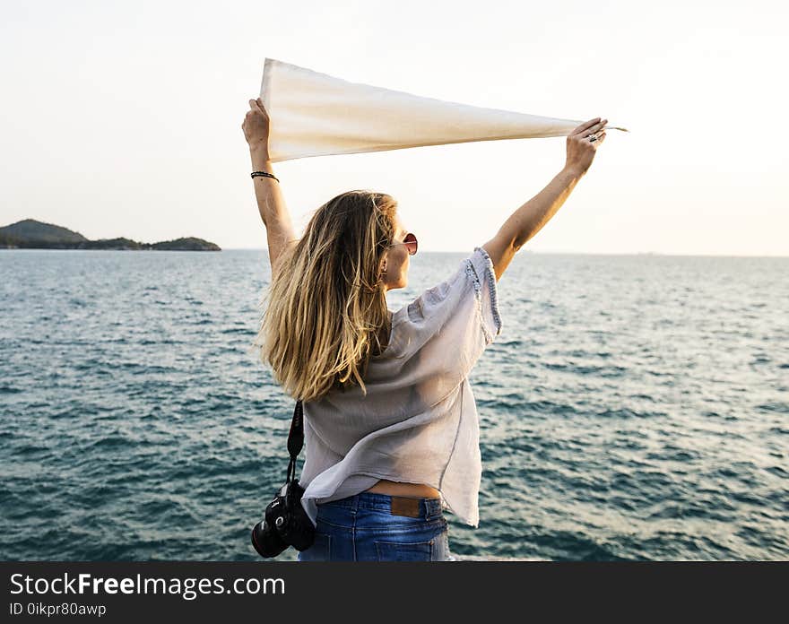 Photo of Woman Wearing White Top, Blue Bottoms and Black Dslr Camera Holding White Textile While Facing the Ocean