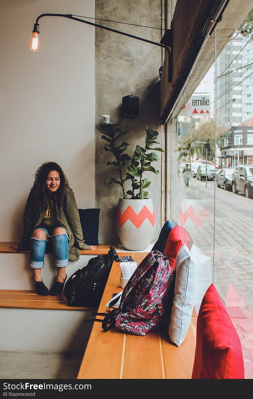 Woman in Gray Zip Up Jacket and Blue Distressed Jeans Sitting on Bench Near Backpack Looking Outside in Room