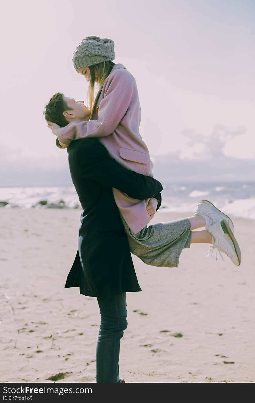 Man in Gray Coat Carrying Woman Wearing Pink Coat in Beach Near Shoreline and Body of Water