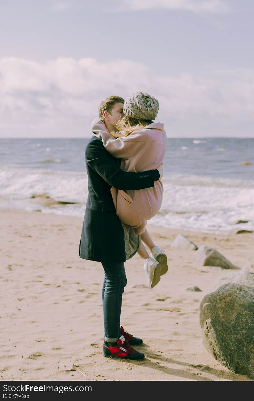 Man in Black Overcoat and Blue Denim Jeans Kissing While Carrying a Woman in Pink Overcoat and Knit Cap on Shore at Daytime