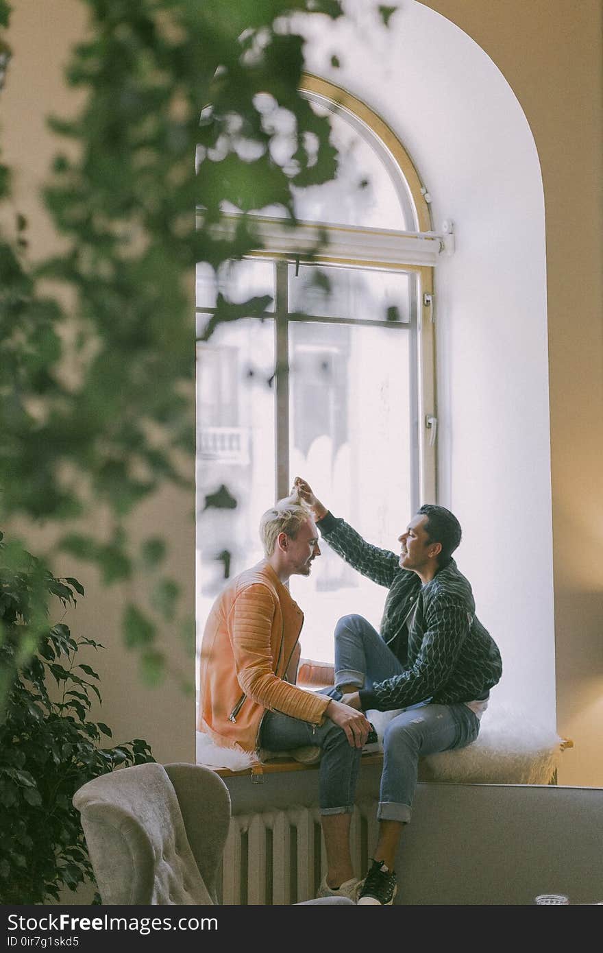 Man Wearing Green Jacket in Front of Man Wearing Orange Zip-up Jacket Sitting on Near the Window Photo