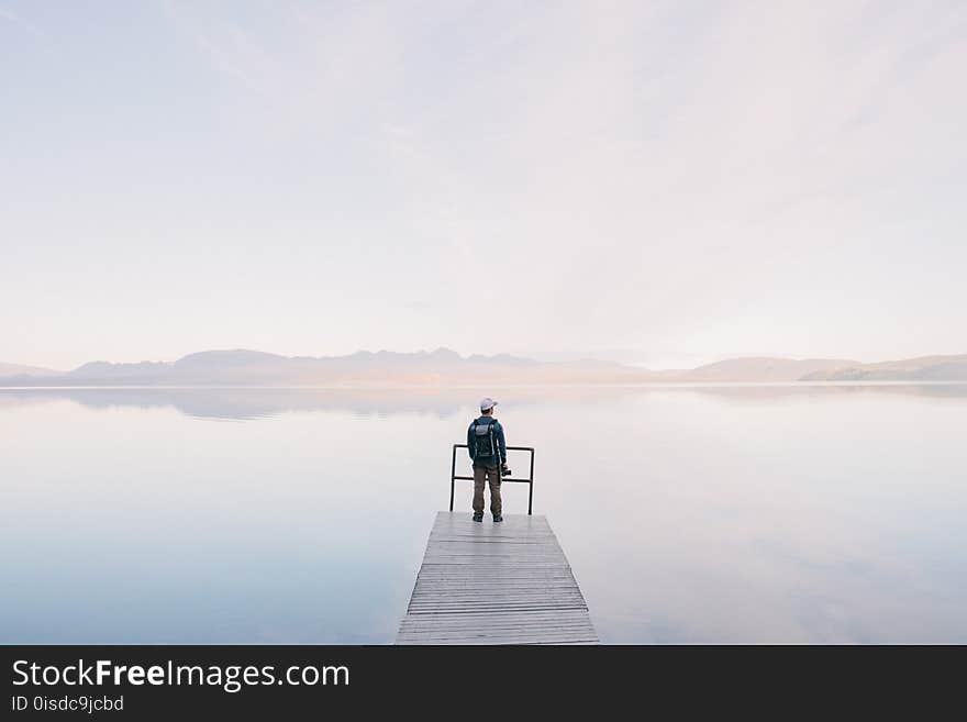 Man Wearing Jacket Standing on Wooden Docks Leading to Body of Water