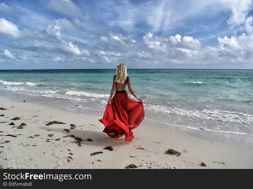 Woman Wearing Red Dress Walking on Seashore Under Blue and White Sky