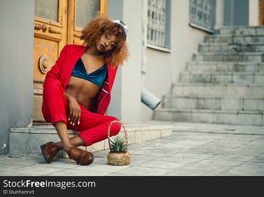 Woman Wearing Red Coat and Pants Sitting Outside Gray Building