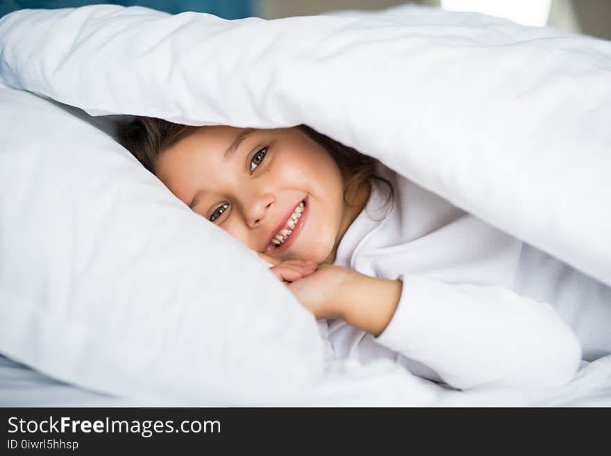 Pretty little girl in sleepwear lying under blanket in the bed at home