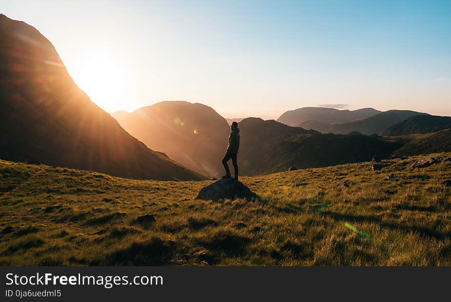 Silhouette Photography of Person Standing on Green Grass in Front of Mountains during Golden Hour