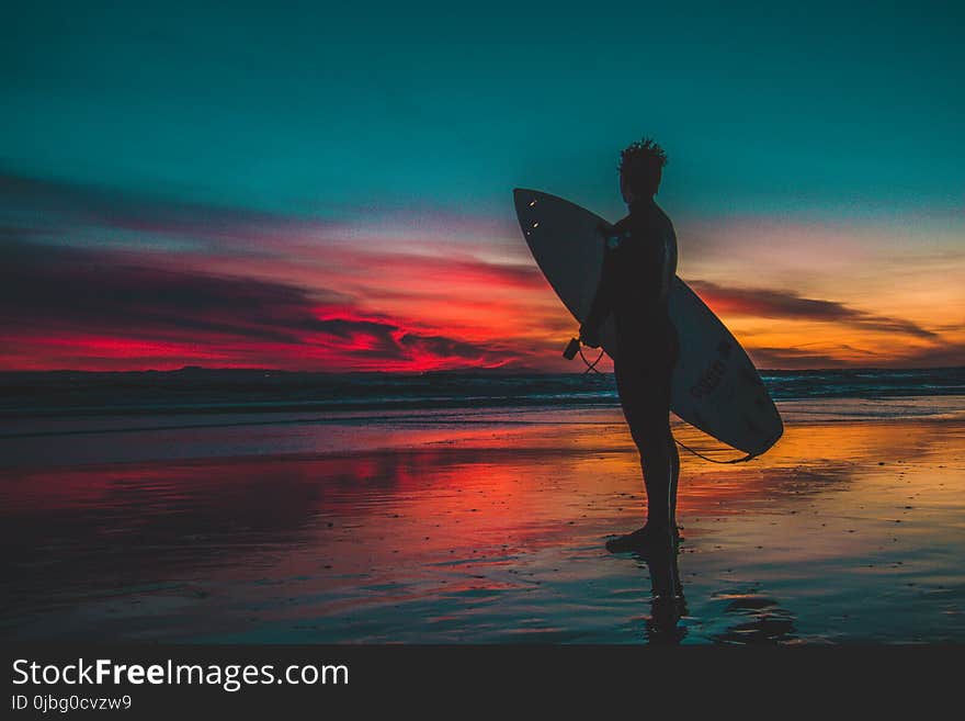 Silhouette of Man in Wet Suit Holding White Surfboard While Standing on Beach during Golden Hour