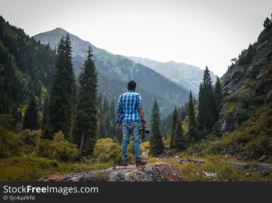 Man in Blue and White Checked Quarter-sleeved Shirt Holding Black Camera Standing on Rock