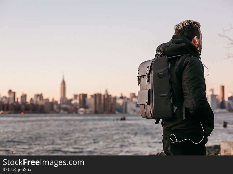 Man Wearing Black Bubble Jacket and Black Leather Backpack Near Bay