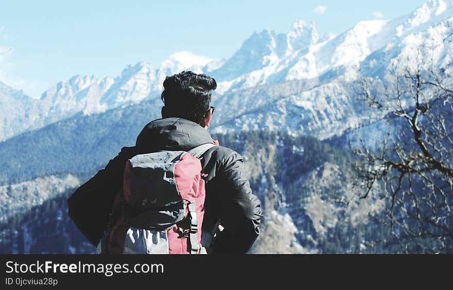 Photography of Man in Black Hooded Jacket and Red Backpack Facing Snow Covered Mountain