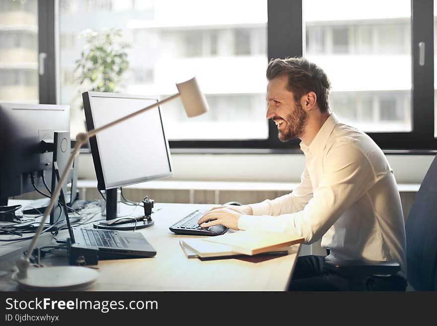 Man in White Dress Shirt Sitting on Black Rolling Chair While Facing Black Computer Set and Smiling