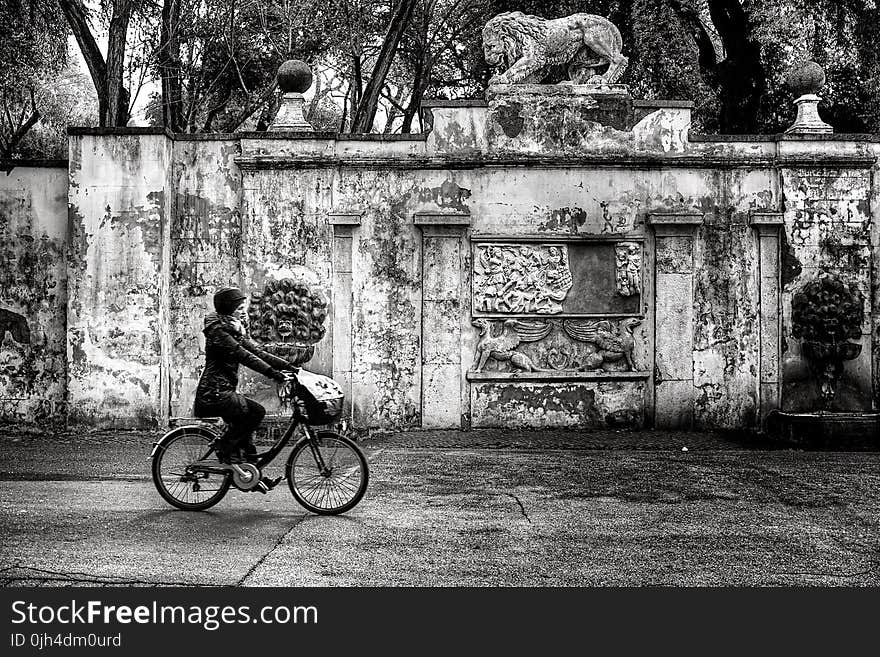 Woman Wearing Jacket and Pants Riding on Bicycle Near Concrete Wall Greyscale Photo