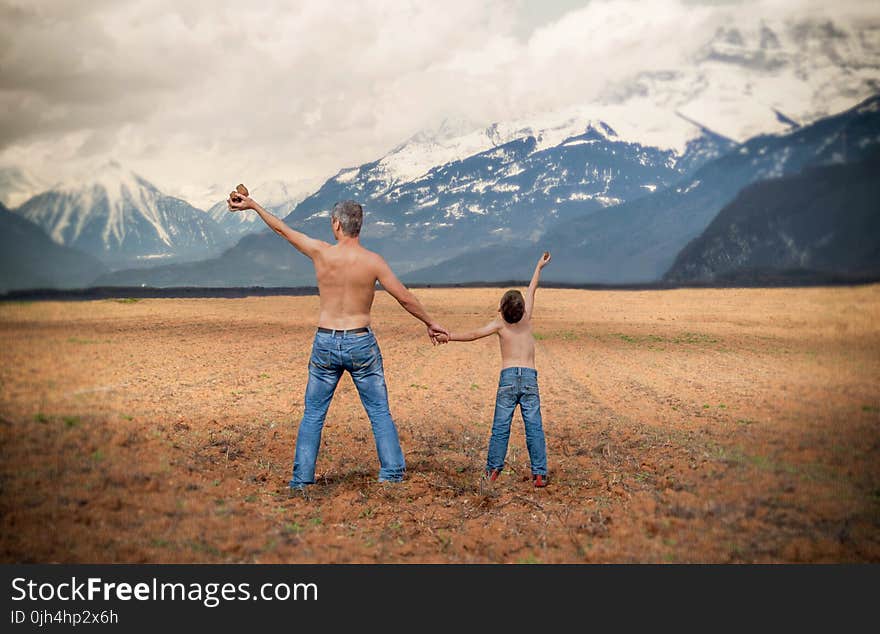 Man and a Boy in Blue Denim Jeans Standing in Brown Open Space Near White and Gray Snowy Mountains during Daytime