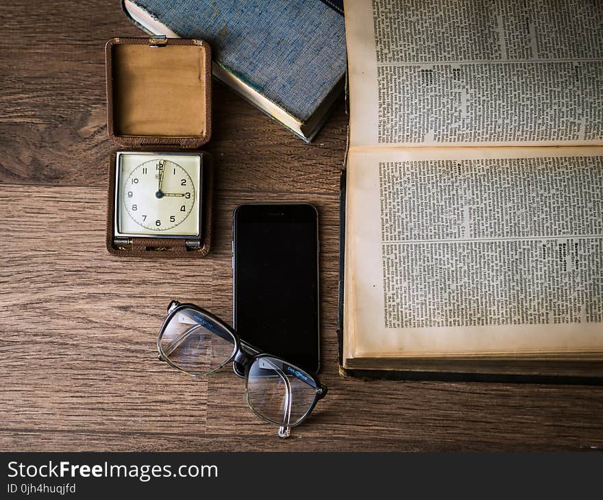 Black Framed Eyeglasses on Top of Black Smartphone Near Brown Square Analog Clock on Brown Wooden Surface