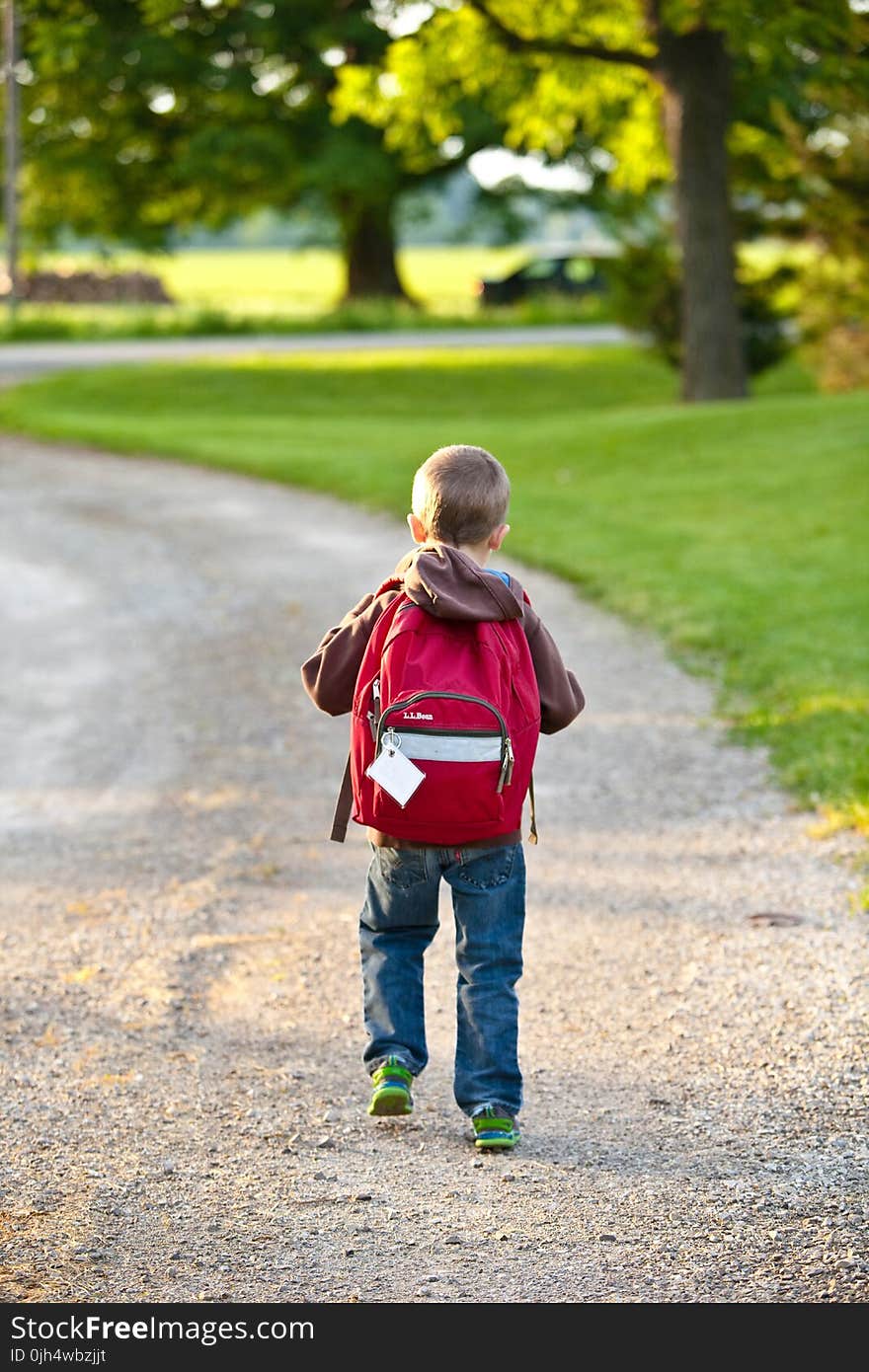 Boy in Brown Hoodie Carrying Red Backpack While Walking on Dirt Road Near Tall Trees