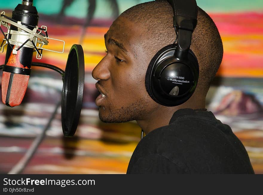 Man in Black Tops Wearing Black Headphones Singing in Front of Black Condenser Microphone