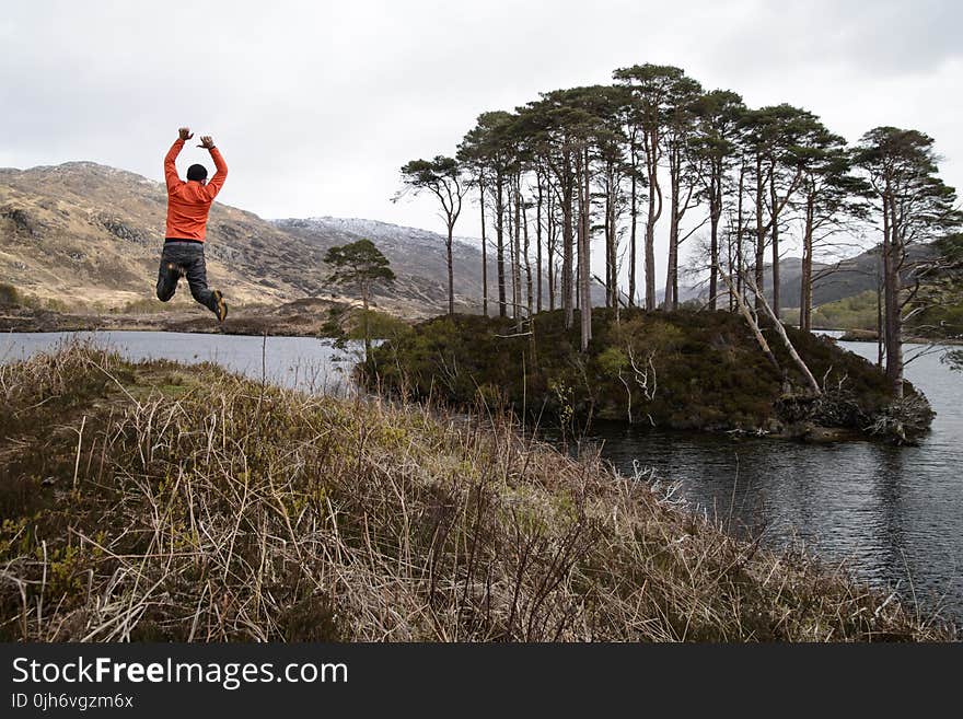 Man in Orange Long-sleeved Shirt Jumping on Lake Near Tall Trees at Daytime