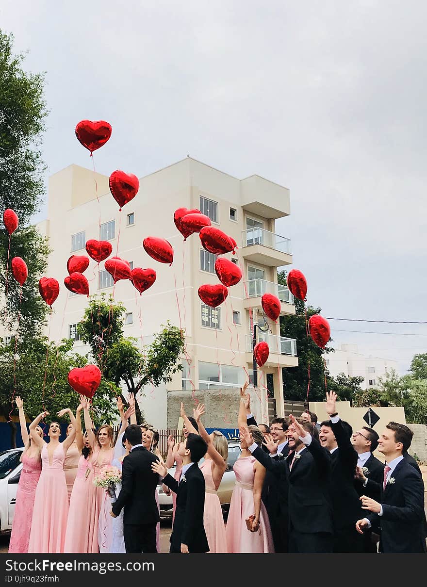 Women Wearing Pink Dresses and Men Wearing Black Suit Jacket and Pants Raising Hands With Red Heart Balloons