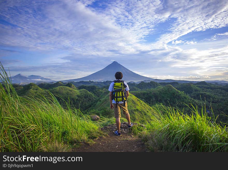 Man Wearing White Shirt, Brown Shorts, and Green Backpack Standing on Hill