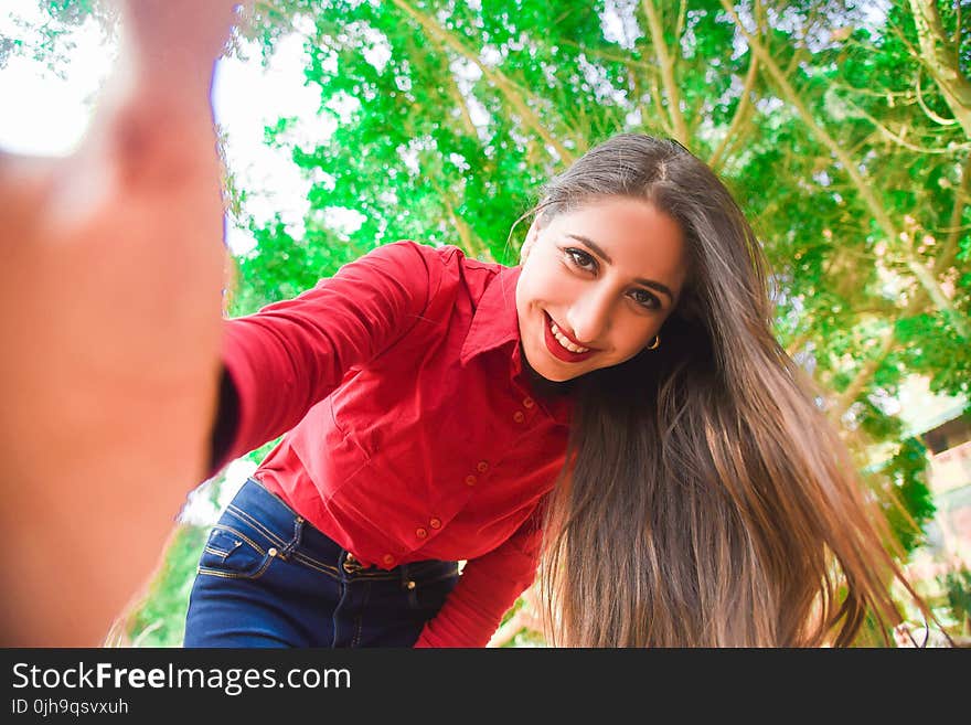 Smiling Woman in Red Shirt and Blue Jeans Taking Selfie Under Green Leaved Tree