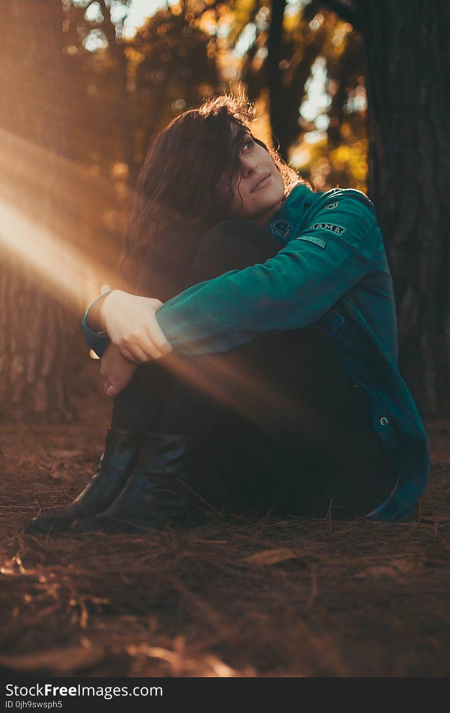 Selective Focus Photography of Woman in Green Jacket and Black Pants Sitting on Field Surrounded With Trees