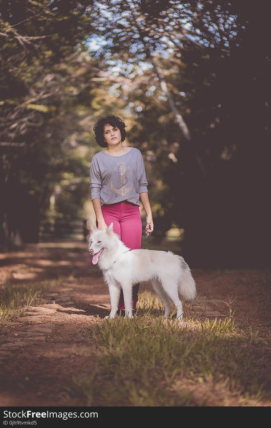 Woman in Purple Long Sleeve Shirt and Pink Jeans Standing Next to White German Spitz With Background of Green Trees during Day