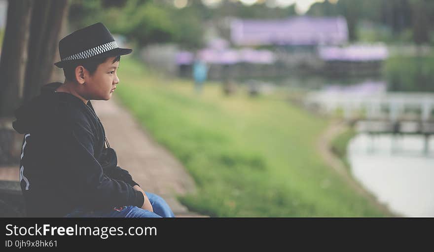 Boy in Black Jacket and Black Fedora Hat Sitting Near Body of Water