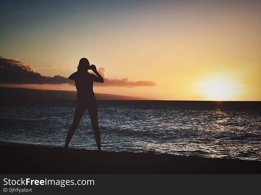 Silhouette of Woman Holding Camera Near Seashore during Golden Hour