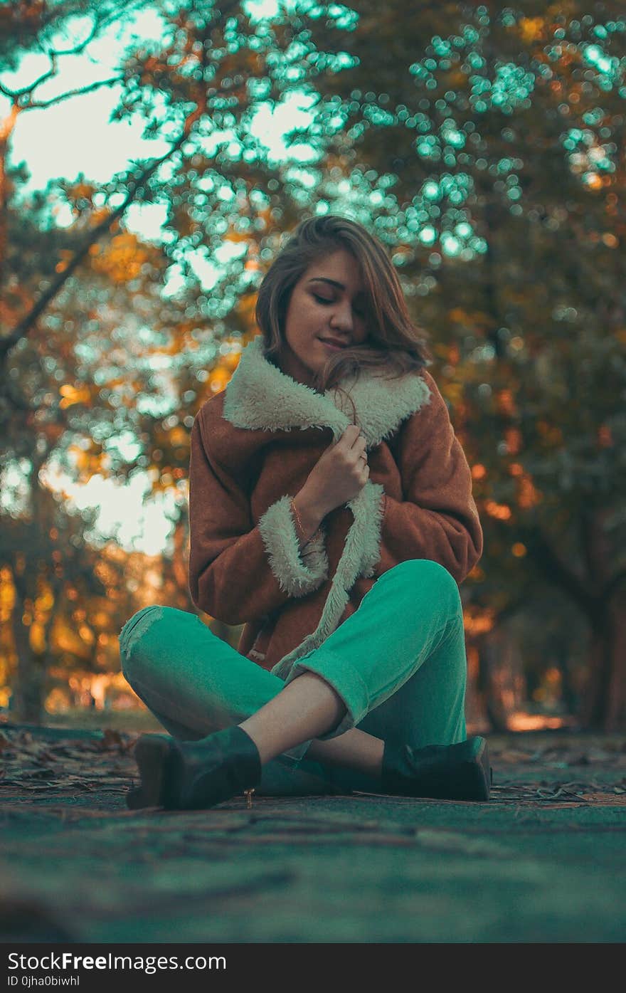 Woman Wearing Brown-and-white Coat and Teal Denim Jeans Sitting on Road Surface