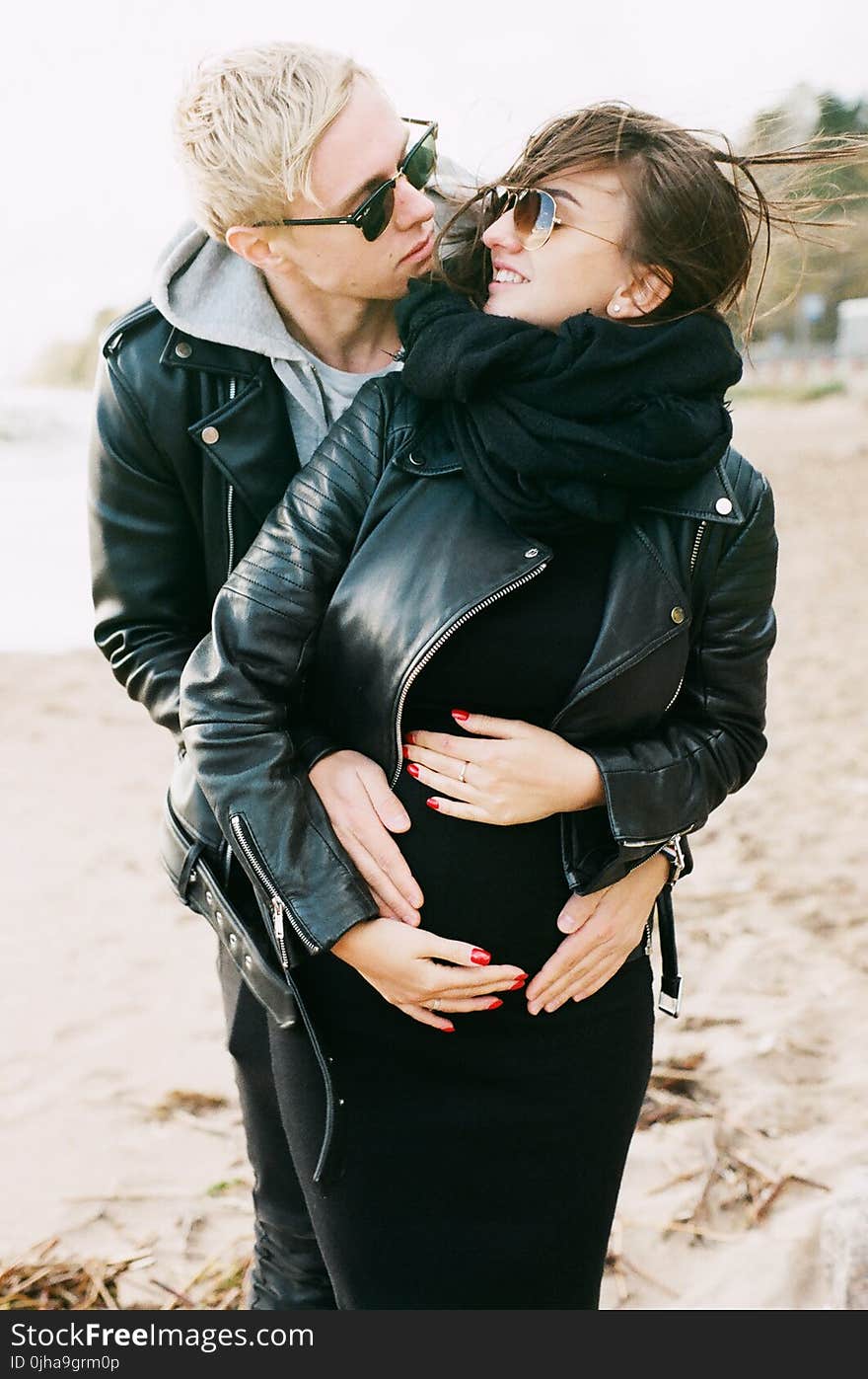 Man in Black Leather Zip Jacket Hugging Woman in Black Leather Zip Jacket and Black Pants Standing on Sand at Daytime