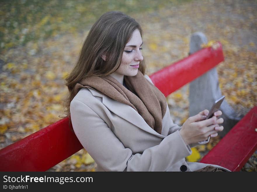 Woman in Beige Coat Holding Smartphone Sitting on Bench