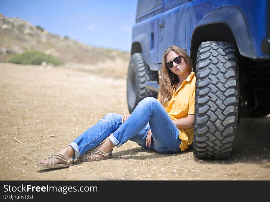 Woman in Yellow Polo Shirt Sitting on Ground Leaning on Blue Vehicle at Daytime