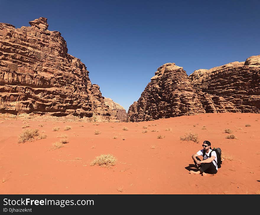 Man Wearing White Shirt and Black Pants Sitting on Soil Behind Rock Formation Mountain