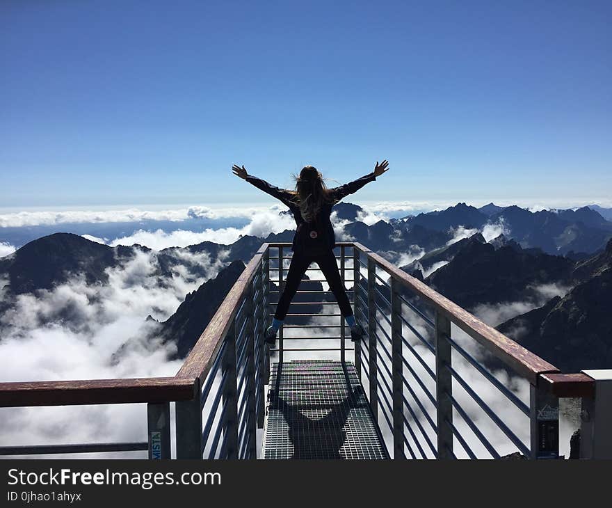 Person Standing on Hand Rails With Arms Wide Open Facing the Mountains and Clouds